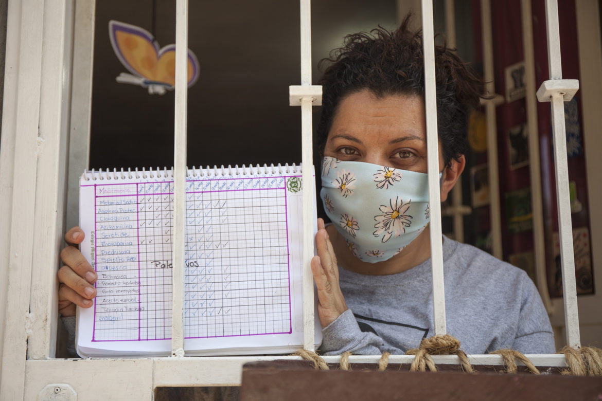 Mary Gloria Fournier, habitante de la colonia Guerrero, tras la reja del espacio donde está en cuarentena, muestra su bitácora de cuidados y dosis de medicamentos para Juan, su compañero de vida, enfermo de COVID19. Foto: César Martínez López.